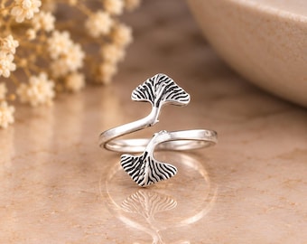 Puede incluir: Un anillo abierto de tono plateado que presenta dos motivos de hojas de ginkgo enfrentados. El metal tiene un acabado oxidado en las áreas hundidas para resaltar los patrones detallados de las venas de las hojas, creando un contraste.