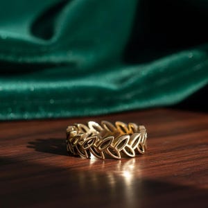 May include: A gold ring featuring a repeating leaf pattern. The ring sits on a dark wooden surface, with a green fabric backdrop. The ring's details are accentuated by the lighting, creating shadows.