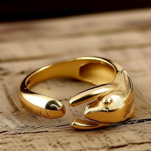 Puede incluir: Un anillo dorado con diseño de conejo. El anillo presenta una cabeza de conejo detallada en un extremo y una banda lisa y ajustable. Las orejas y la cara del conejo están claramente definidas, creando una joya única y llamativa.