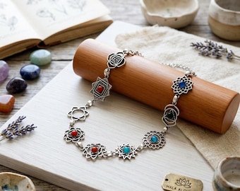 May include: A silver-toned metal chain necklace featuring seven distinct geometric charms, each inlaid with a small round cabochon stone in red, blue, or green. The necklace is displayed on a cylindrical wooden stand next to a metal tag with the text MALA ALCHEMY.