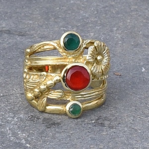 May include: A gold-colored ring with a multi-band design. The ring features three gemstones: a central red stone and two green stones. Floral and leaf-like details adorn the ring's bands, creating an ornate appearance.