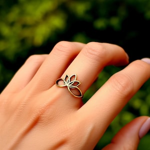 May include: A gold-coloured ring shaped like a lotus flower. The ring is worn on a finger, with the flower design facing upwards. The ring's design is delicate and open, with the petals clearly defined.