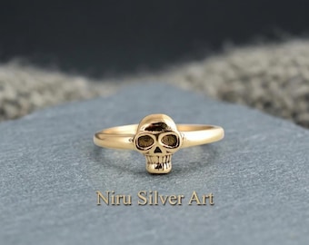 Peut inclure: Une bague dorée avec un motif de crâne central. L'anneau est lisse et circulaire, se connectant au design détaillé du crâne sur le devant. Le texte "Niru Silver Art" est imprimé dans une police claire sous la bague sur la surface.