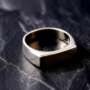 Può includere: Un anello chevalier in argento con una superficie piatta e rettangolare e una finitura lucida. L'anello è posto su uno sfondo scuro e strutturato, che ne evidenzia il design elegante. Questo gioiello è adatto all'uso quotidiano.