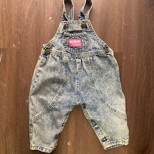 Pode incluir: Macac&atilde;o de ganga azul claro com um patch de logotipo vermelho OshKosh B'Gosh. O macac&atilde;o tem al&ccedil;as ajust&aacute;veis e fechos de press&atilde;o.