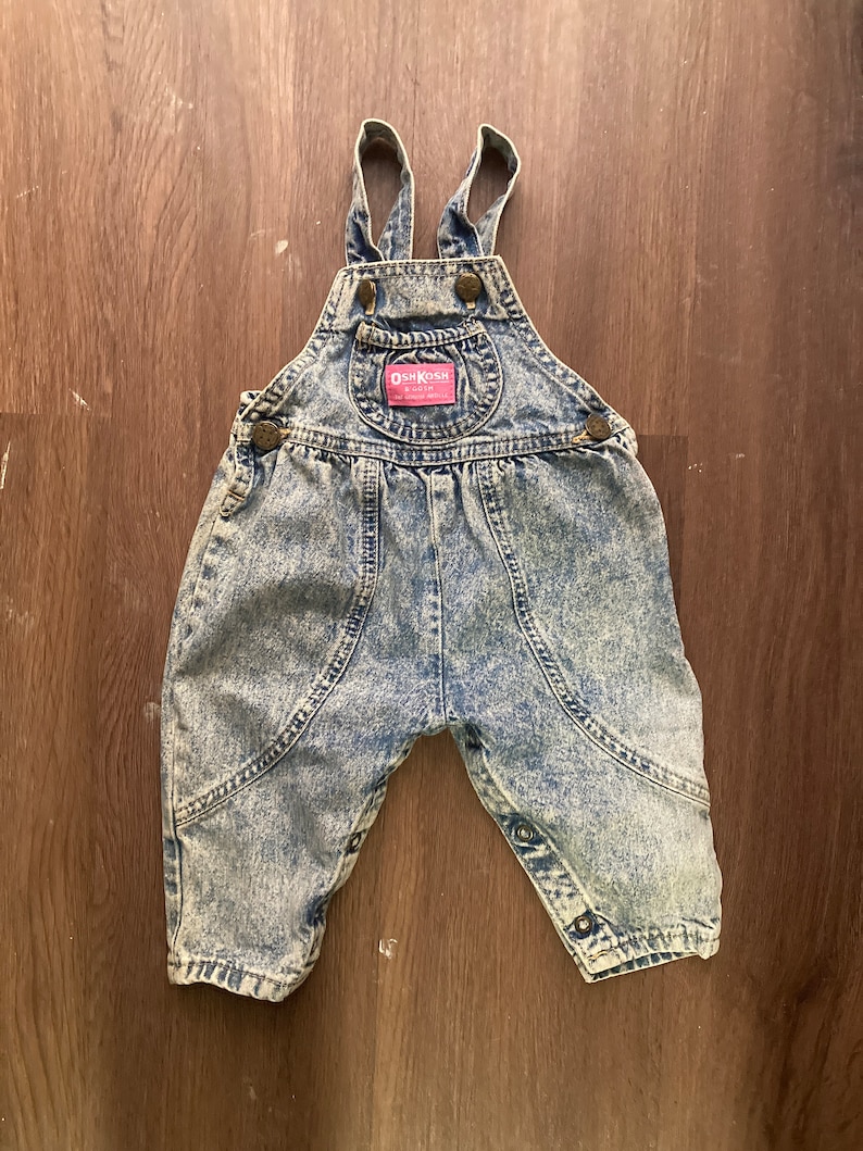 Pode incluir: Macac&atilde;o de ganga azul claro com um patch de logotipo vermelho OshKosh B'Gosh. O macac&atilde;o tem al&ccedil;as ajust&aacute;veis e fechos de press&atilde;o.