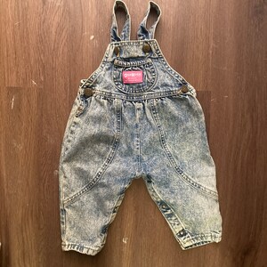 Pode incluir: Macac&atilde;o de ganga azul com um acabamento de lavagem &aacute;cida clara. O macac&atilde;o tem al&ccedil;as ajust&aacute;veis e um remendo com o logotipo OshKosh B'Gosh.