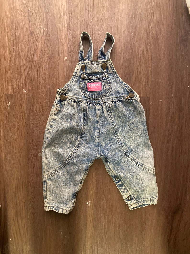 Pode incluir: Macac&atilde;o de ganga azul com um acabamento de lavagem &aacute;cida clara. O macac&atilde;o tem al&ccedil;as ajust&aacute;veis e um remendo com o logotipo OshKosh B'Gosh.