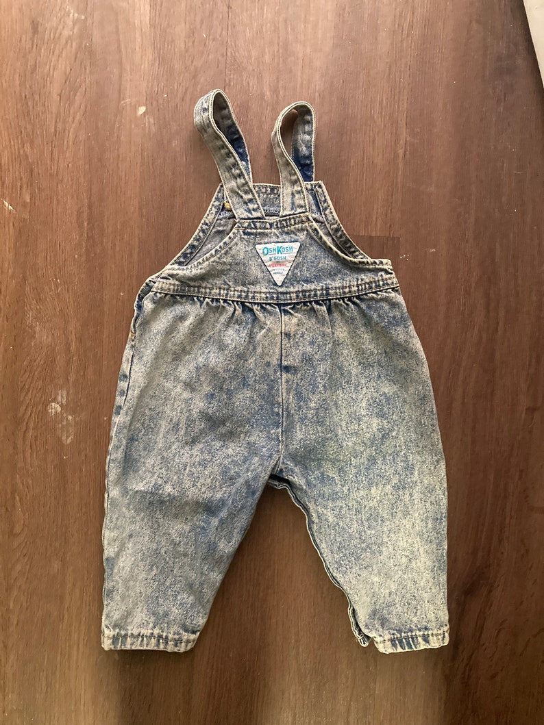 Pode incluir: Macac&atilde;o de ganga azul claro lavado com &aacute;cido com al&ccedil;as ajust&aacute;veis e um patch com o logotipo OshKosh B'Gosh.