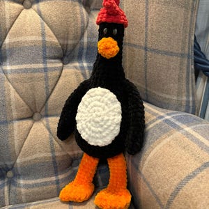 Puede incluir: Un pingüino de crochet negro y blanco con un gorro de crochet rojo. El pingüino tiene pies de crochet naranja y está sentado en una silla a cuadros.