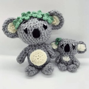 May include: Two crocheted koala bears, one larger than the other. The larger koala has a green leaf crown and a white belly patch. The smaller koala has a single green leaf on its head and a white belly patch.