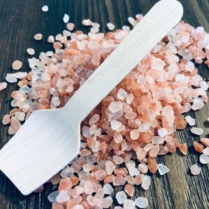 May include: A wooden scoop rests on a pile of pink Himalayan salt crystals. The salt crystals are various sizes and shades of pink, with some white crystals mixed in. The scoop is light-colored and has a rectangular bowl.