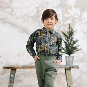 May include: A young person wearing a green and yellow patterned button-down shirt, green pants, and black shoes. The person is standing in front of a white wall with a small potted tree behind them.