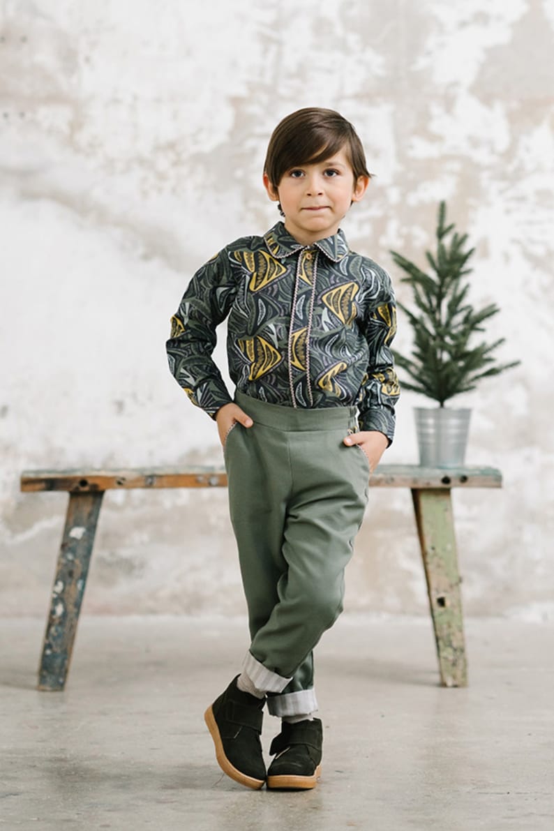 May include: A young person wearing a green and yellow patterned button-down shirt, green pants, and black shoes. The person is standing in front of a white wall with a small potted tree behind them.