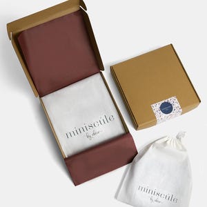 May include: A brown cardboard box with a red tissue paper lining and the text "miniscule by elora" printed on the inside. The box is open and a white drawstring bag with the same text is visible in the foreground.