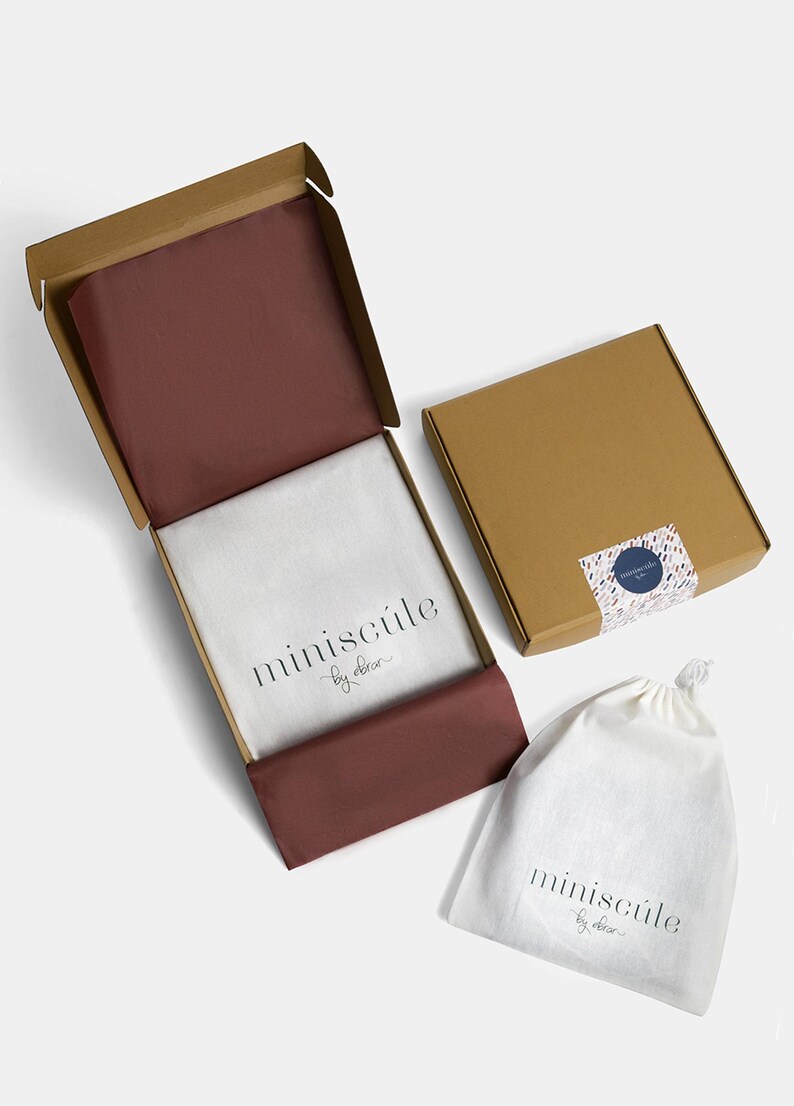 May include: A brown cardboard box with a red tissue paper lining and the text "miniscule by elora" printed on the inside. The box is open and a white drawstring bag with the same text is visible in the foreground.