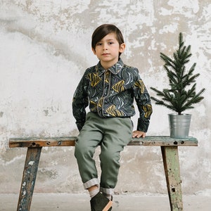 May include: A young person wearing a green and yellow patterned shirt, green pants, and dark green shoes sits on a wooden bench. A small potted evergreen tree is behind the bench.