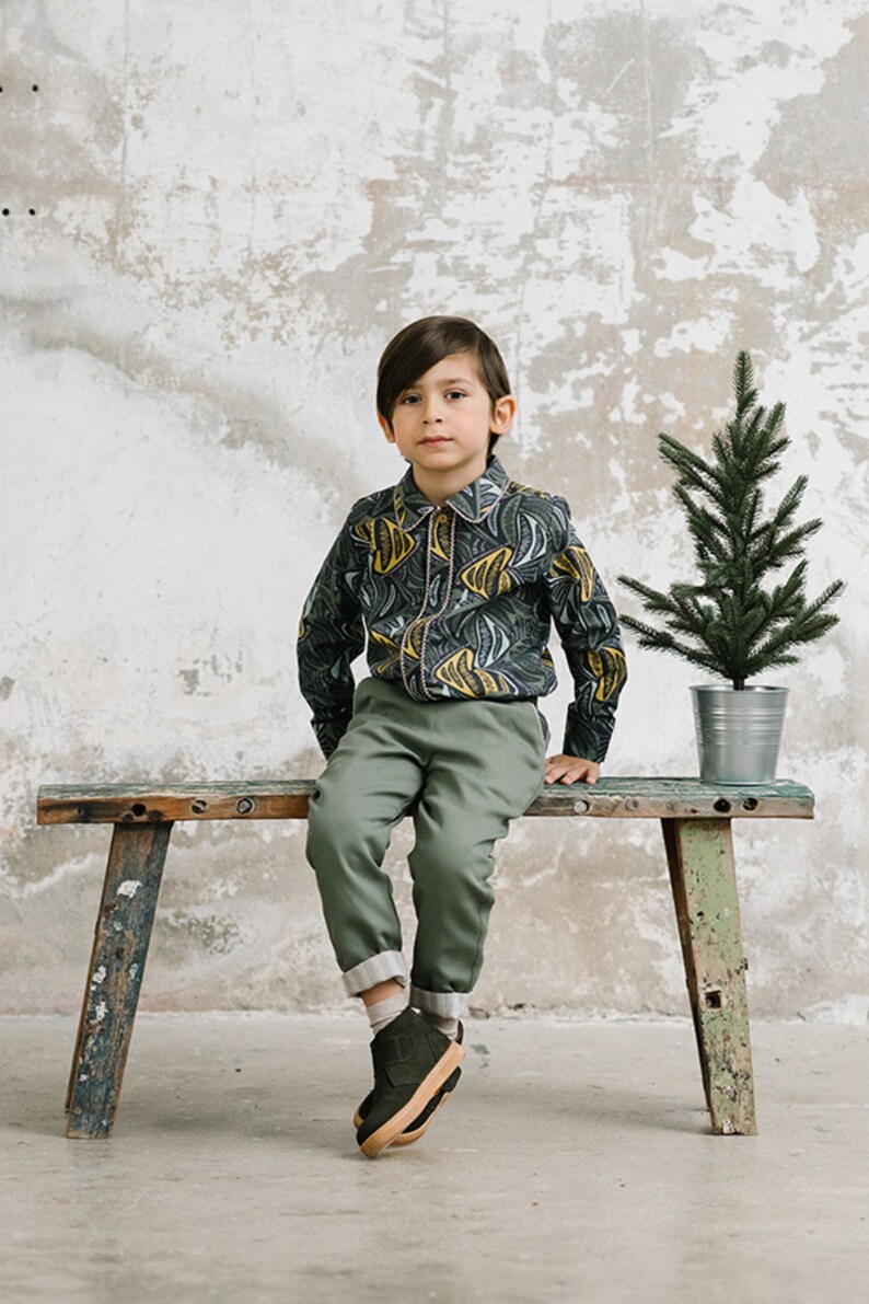 May include: A young person wearing a green and yellow patterned shirt, green pants, and dark green shoes sits on a wooden bench. A small potted evergreen tree is behind the bench.