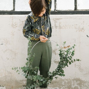 May include: A child wearing a green and yellow patterned shirt, green pants, and black shoes holds a eucalyptus wreath.