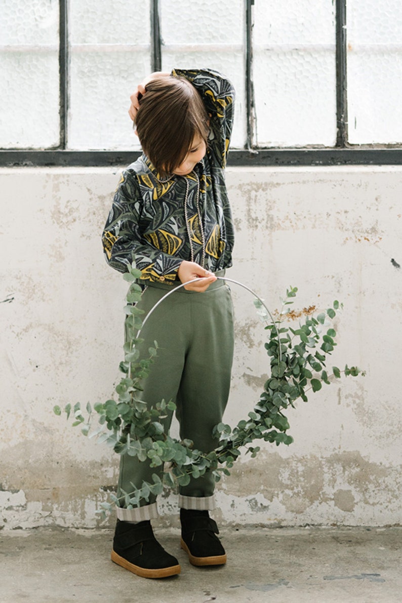 May include: A child wearing a green and yellow patterned shirt, green pants, and black shoes holds a eucalyptus wreath.