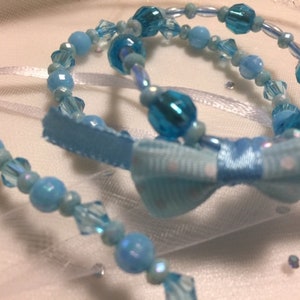 May include: Two blue beaded bracelets with a light blue bow. The bracelets are made of small, round beads and crystal beads.
