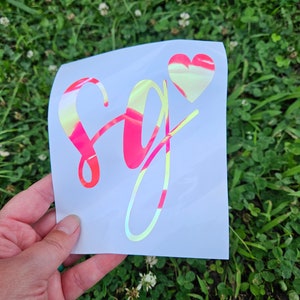 May include: A white vinyl sticker with a holographic finish. The sticker features the letters "SG" in a pink and yellow script font with a small pink heart above the letters.