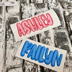 May include: Two white vinyl stickers with names written in bold, colorful letters. The top sticker has the name "ASHLEY" in pink letters. The bottom sticker has the name "PAILYNN" in blue letters.