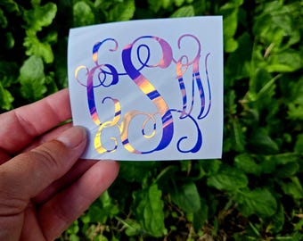 May include: A monogram sticker with a gradient design in shades of purple, blue, and gold. The sticker features three intertwined letters, creating a personalized and decorative element. The sticker is held against a backdrop of green foliage.