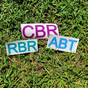 Puede incluir: Tres pegatinas blancas con las palabras "CBR", "RBR" y "ABT" escritas en letras azules y rosas. Las pegatinas están sobre una superficie de hierba verde.
