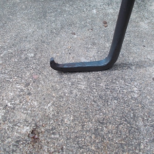 May include: Close-up of a black metal hook with a sharp point. The hook is resting on a gray concrete surface.