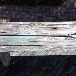 May include: A hand-forged metal fire poker with a hook on one end and a looped handle on the other. The shaft has a twisted design near the handle. The tool is silver and rests on a weathered wooden surface.