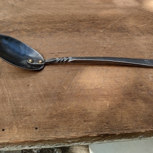 May include: A hand-forged metal spoon with a twisted handle and two brass accents. The spoon has a rounded bowl and a loop handle.