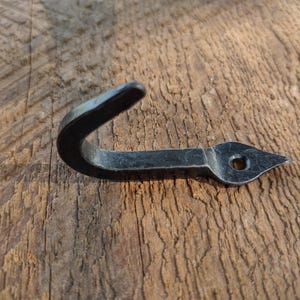 May include: A hand-forged, black metal hook with a curved top and a pointed end with a hole. The hook is designed for hanging items and is set against a rustic wooden background.
