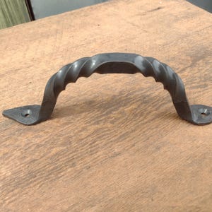 May include: A dark gray, hand-forged metal handle with a twisted design. The handle has a curved shape with two flat mounting plates, each with a pre-drilled hole for screws. This rustic hardware is suitable for cabinets, drawers, or doors.