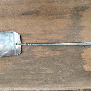 May include: A handcrafted metal spatula with a square head and a long, twisted handle. The spatula is made of silver-colored metal and features a loop at the end of the handle for hanging. The spatula is approximately 20 inches long.