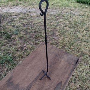 May include: A handcrafted, black metal fire poker with a looped handle and a twisted shaft. The poker stands upright on a wooden surface, with a cross-shaped base for stability. The background is a grassy area.