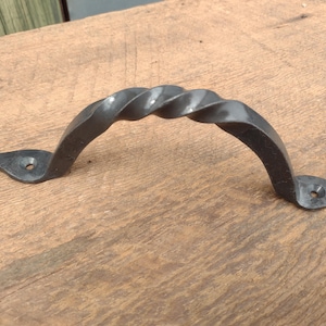May include: A dark gray, hand-forged metal handle with a twisted design. The handle has a curved shape with decorative, leaf-shaped mounting plates on each end. The handle is designed for use on doors, drawers, or cabinets.