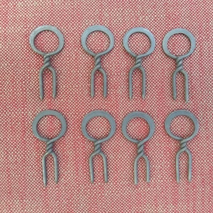 May include: Eight silver metal skewers with a twisted handle and a circular loop at the top. The skewers are arranged in two rows of four on a red textured surface.
