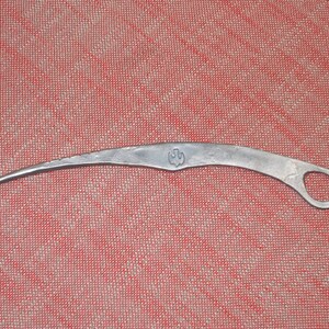 May include: A silver metal tool with a curved blade and a small ring at the end. The tool has a stylized bird design on the blade.
