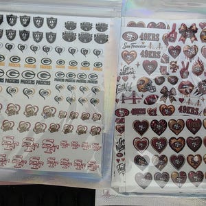 May include: Two clear plastic pouches containing sheets of adhesive decals. The left sheet features Las Vegas Raiders and Green Bay Packers logos and heart-shaped designs. The right sheet displays San Francisco 49ers logos, hearts, footballs, and text graphics.