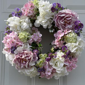 May include: A circular floral wreath composed of various flowers in shades of white, pink, purple, and green. The wreath features large peony-like blooms and smaller hydrangea and other flower varieties, creating a full and textured appearance. The wreath is likely for door or wall decor.