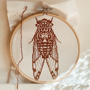 May include: A brown embroidered cicada on white fabric in a wooden embroidery hoop.