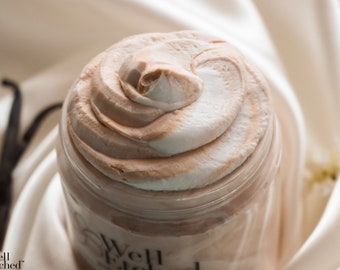 May include: A jar of whipped body butter with a swirl of light brown and white cream. The jar is clear, and the label reads "Well Etched". Vanilla beans and white flowers are in the background.
