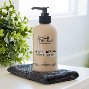 May include: A beige bottle of moisturizing hand soap with a black pump, labeled "Well Etched." The bottle sits on a dark gray washcloth. Green leafy plants are in the background. The soap is 8 oz.
