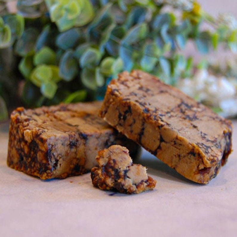 May include: Two rectangular blocks of a light brown substance with dark brown marbling. A small piece is broken off, revealing the interior texture. The blocks are on a light surface, with green foliage in the background.