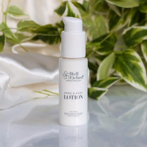 May include: A white hand and body lotion bottle with a pump dispenser. The bottle has the text "Well Etched" and "Hand & Body LOTION". The background includes white fabric and green and white leaves.