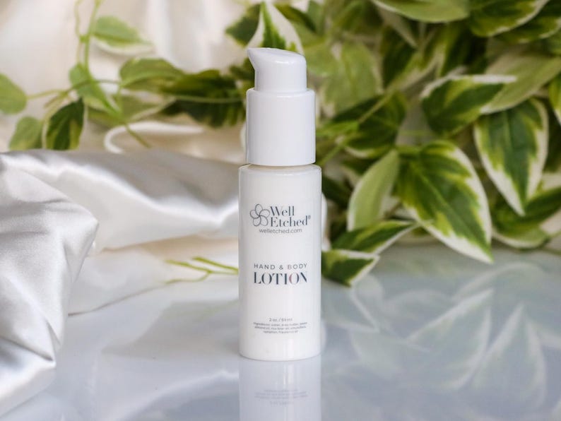 May include: A white hand and body lotion bottle with a pump dispenser. The bottle has the text "Well Etched" and "Hand & Body LOTION". The background includes white fabric and green and white leaves.