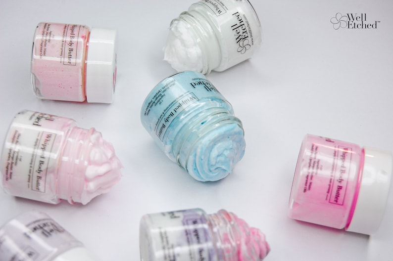 Pu&ograve; includere: Sei piccoli barattoli di vetro pieni di burro corporeo montato. I barattoli sono etichettati "Whipped Body Butter" e hanno un motivo floreale sul coperchio. I barattoli sono su una superficie bianca.