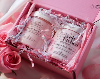 May include: A pink gift box with white tissue paper and two pink jars of body care products. The jars are labeled "Well Etched" and contain "Sugar Scrub" and "Whipped Body Butter".