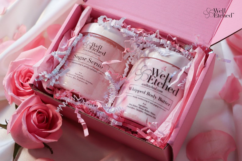 May include: A pink gift box with white tissue paper and two pink jars of body care products. The jars are labeled "Well Etched" and contain "Sugar Scrub" and "Whipped Body Butter".